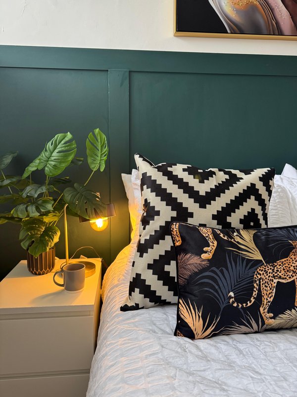 Bedroom with green panelled headboard, bed with patterned pillows, bedside table with plant and lamp