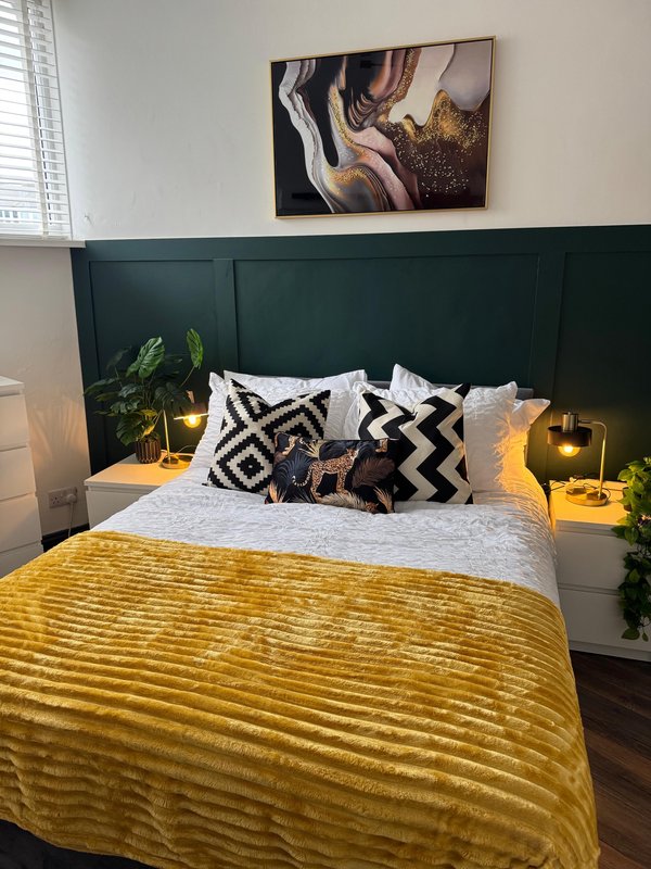 Modern bedroom with dark green headboard, yellow bedding, patterned cushions and wall art