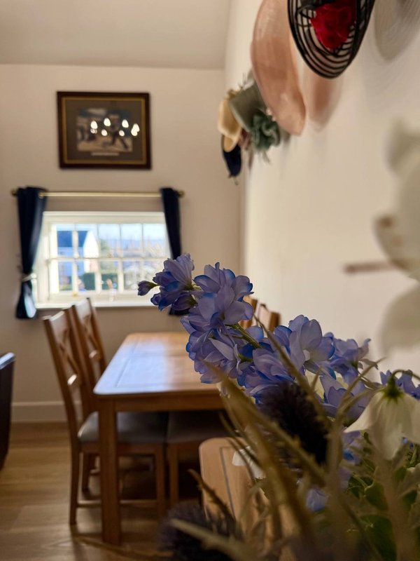 Dining room with wooden table, chairs, window with blue curtains, and purple flowers in foreground