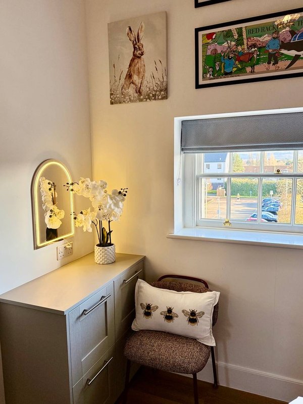 Bedroom corner with white desk, mirror, flowering plant, cushioned chair and framed wall art