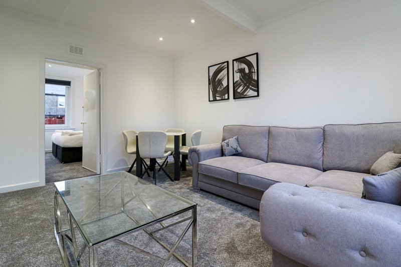 Modern living room with grey sofa, glass coffee table, and dining area visible through doorway