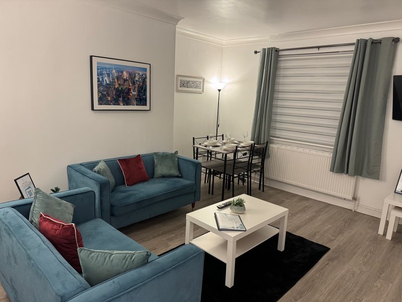 Modern living room with two blue sofas, white coffee table, dining area, and large windows with grey curtains