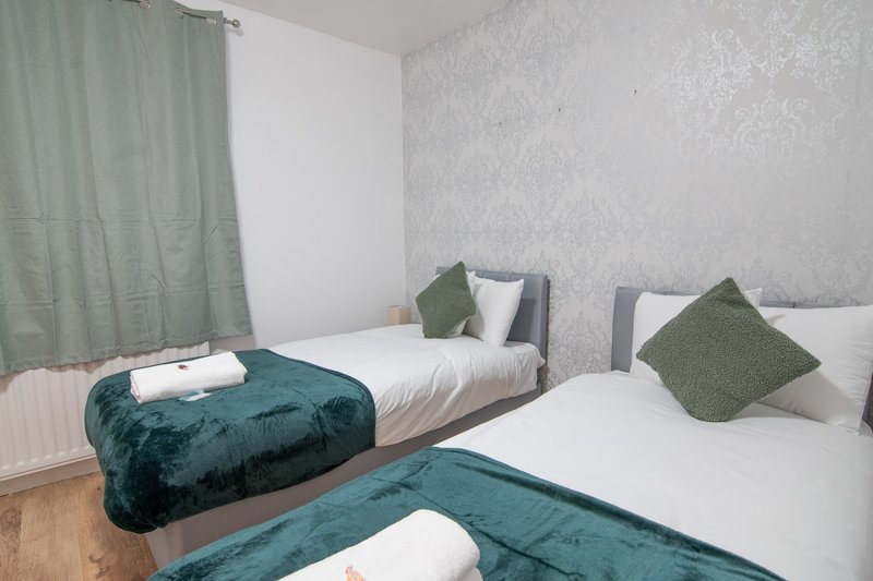 Twin bedroom with two single beds, teal velvet bench, green cushions and pale wallpaper