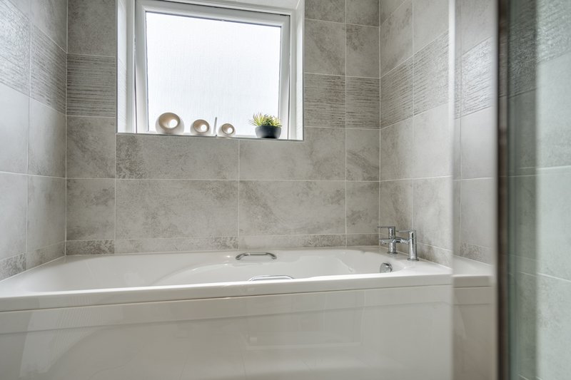 Modern bathroom with white bathtub, grey tile walls, and window with shelf