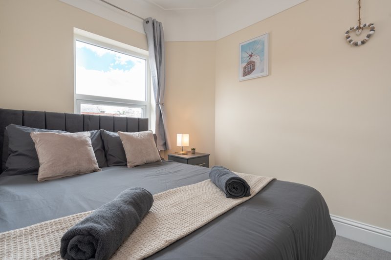 Bright bedroom with double bed, grey headboard, cushions, rolled towels and bedside lamp