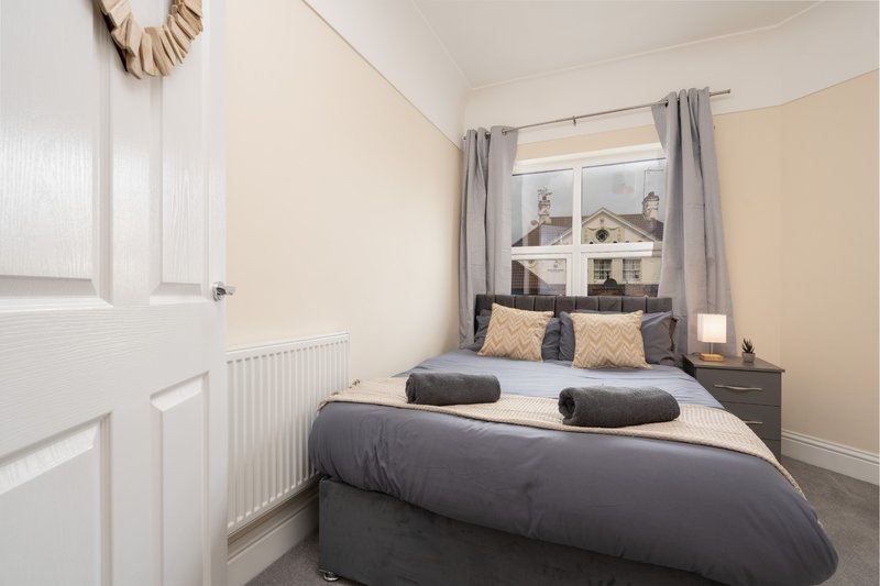 Bedroom with double bed, dark bedding, cushions, radiator and window with grey curtains