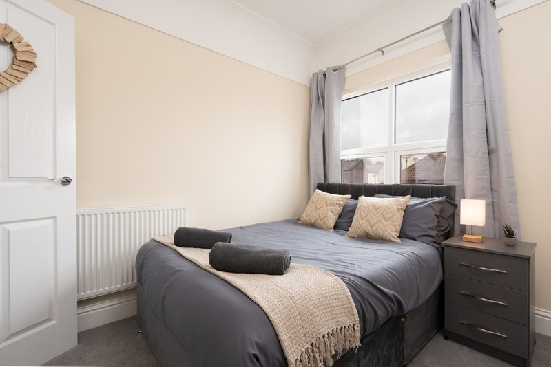 Bedroom with double bed, dark bedding, cream and black cushions, window with grey curtains, radiator and bedside table