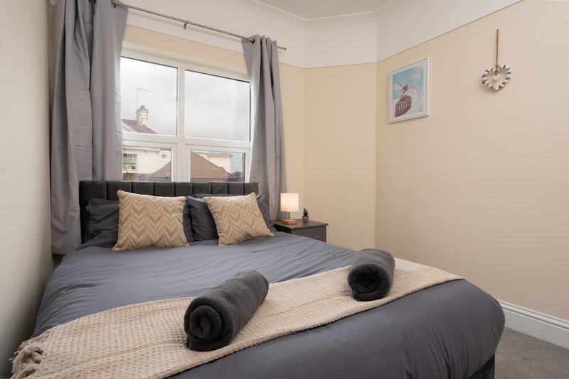 Bright bedroom with double bed, patterned cushions, dark bedding and window with grey curtains