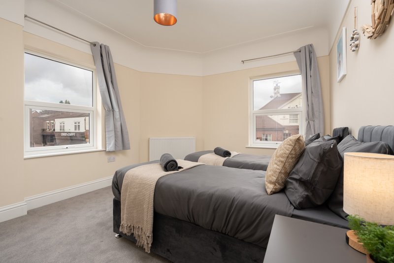 Bright bedroom with twin beds, grey bedding, cushions, and views of neighbouring houses through windows