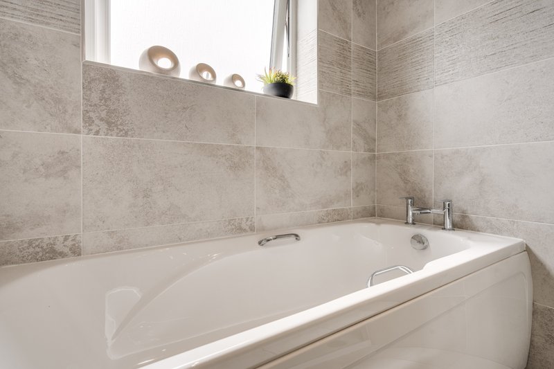 Modern bathroom with white bathtub, beige tile walls, and window with decorative items on sill