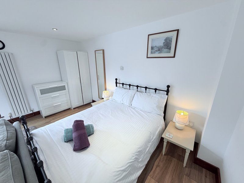 Bright bedroom with double bed, black metal frame, white bedding, wooden floor, wardrobe and bedside lamp