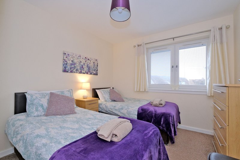 Bright bedroom with two single beds, purple bedding, wooden furniture and window with curtains