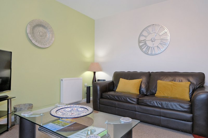 Living room with dark leather sofa, yellow cushions, glass coffee table, wall clocks and yellow accent wall