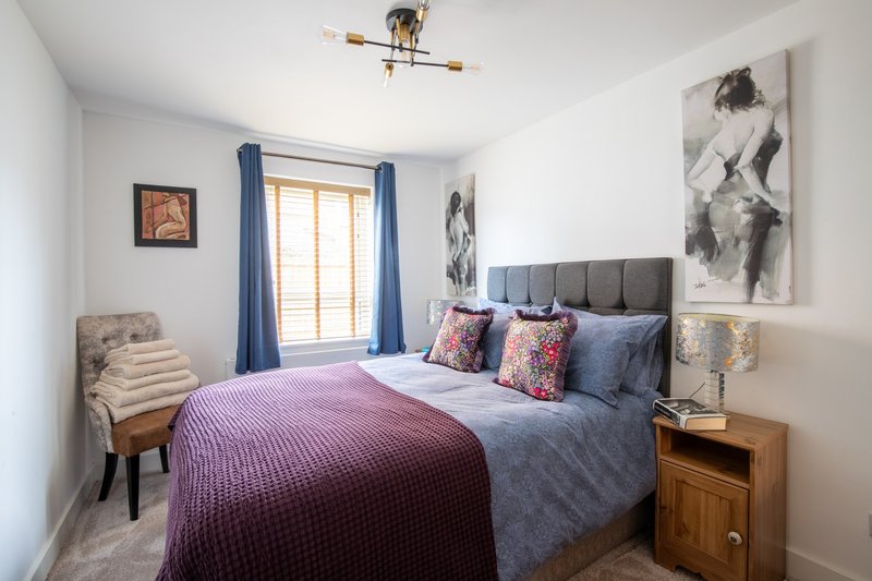 Bedroom with double bed, purple bedding, wooden nightstands, blue curtains and wall art