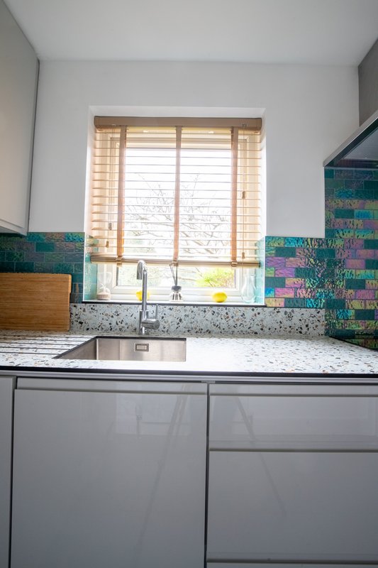 Modern kitchen with white cabinetry, speckled countertop, teal tile splashback and window overlooking garden