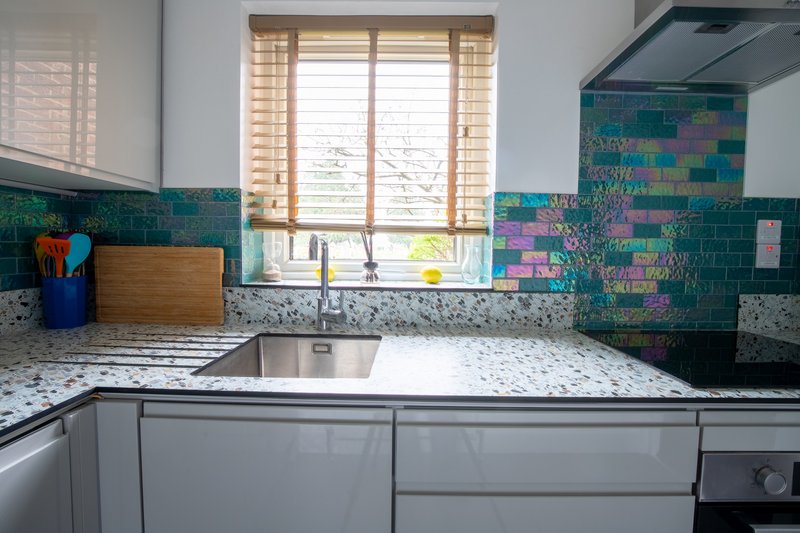 Modern kitchen with colorful teal tile backsplash, white countertops, and window overlooking garden