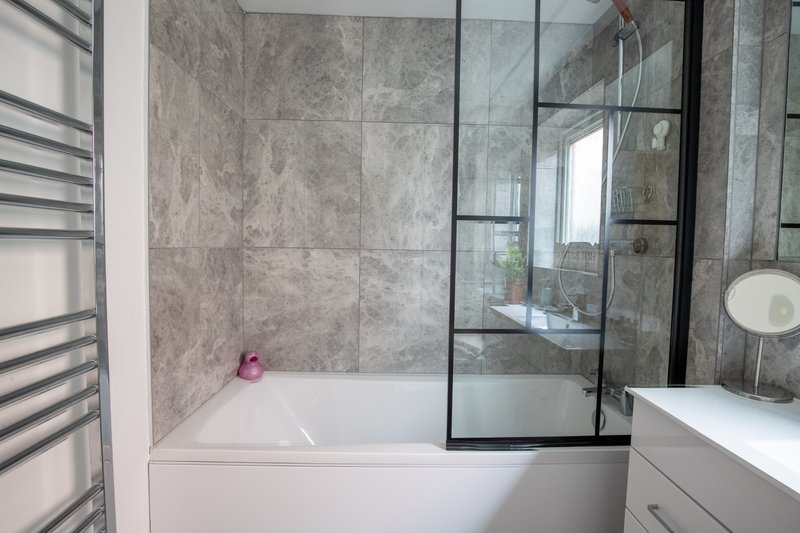 Modern bathroom with white bathtub, grey tile surround, glass shower screen and towel rail