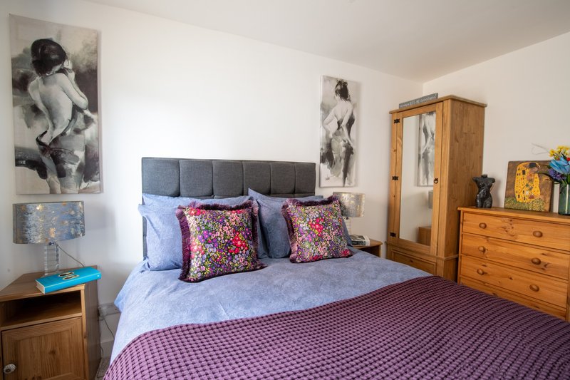 Bedroom with double bed, grey upholstered headboard, decorative cushions, wooden furniture and wall art