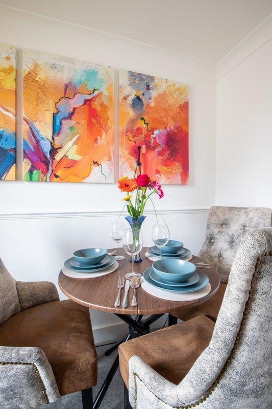 Dining area with round wooden table, place settings, fresh flowers, and upholstered chairs beneath colorful abstract artwork