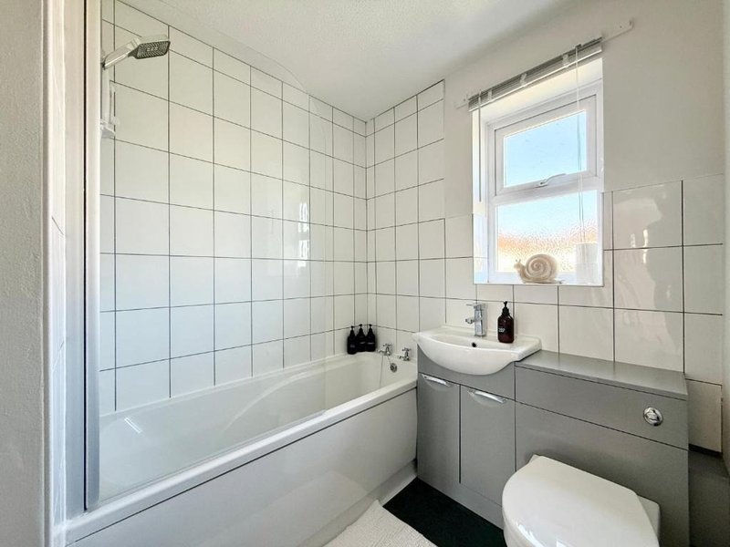 Clean bathroom with white tile walls, bathtub with shower screen, sink vanity, toilet and window
