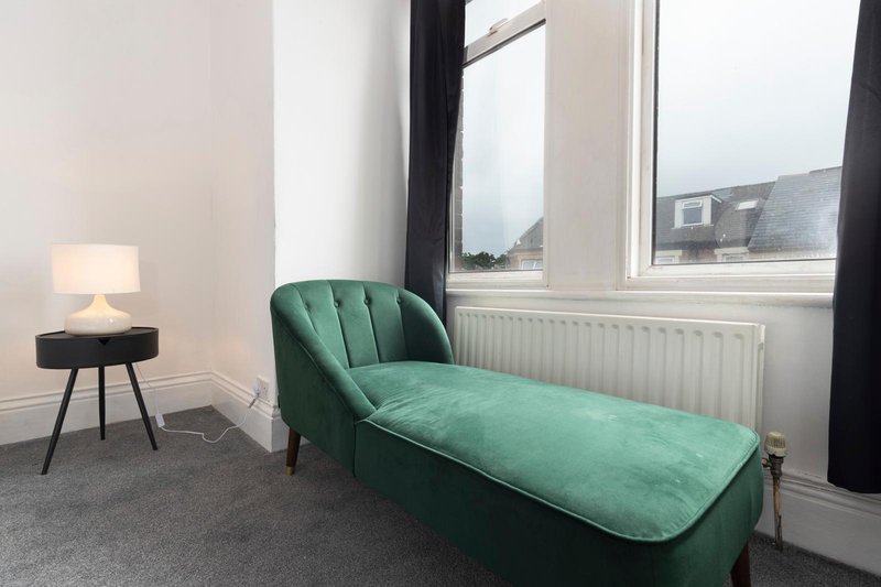 Modern bedroom with green chaise lounge, side table lamp, and large window overlooking street