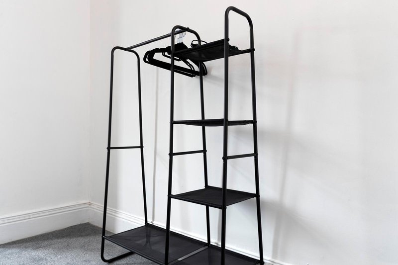Black metal clothing rack with shelves and hangers in corner of white room