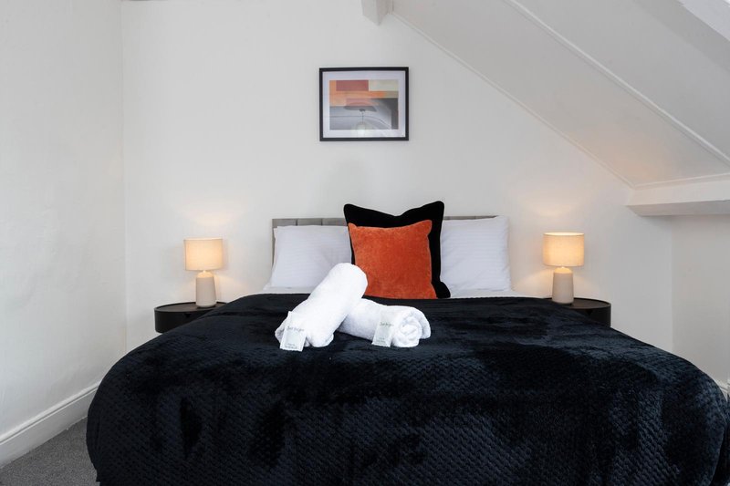 Bedroom with double bed, black bedding, rolled towels, orange cushion and bedside lamps