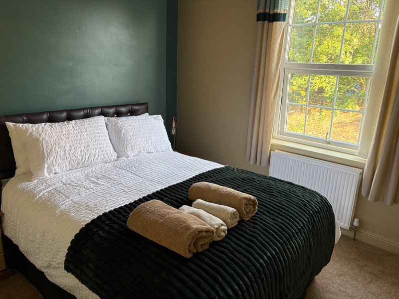 Bedroom with double bed, white pillows, black throw blanket, rolled towels, and large window with garden view