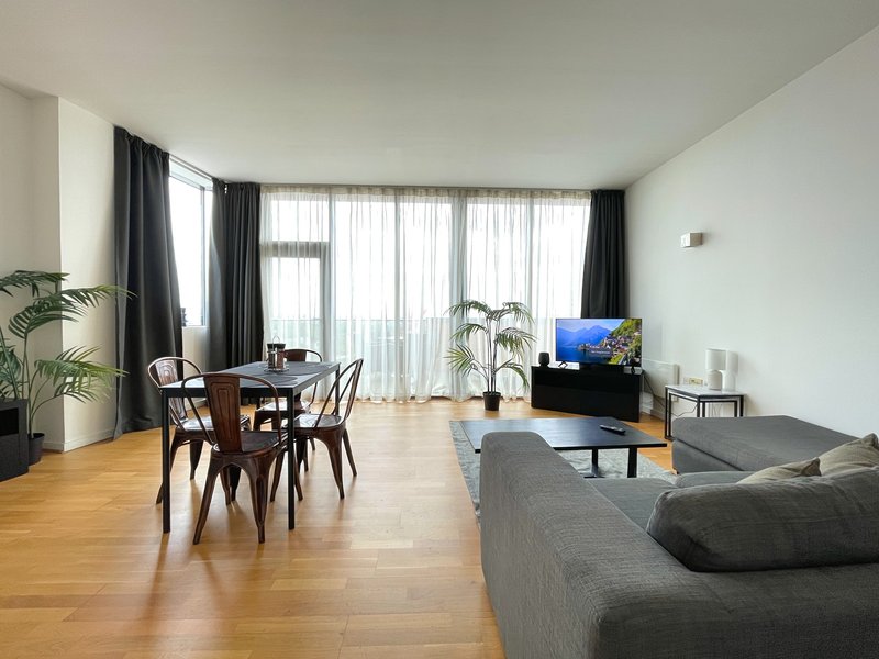 Open-plan living room with dining table, gray sofa, wooden floor and large windows with curtains