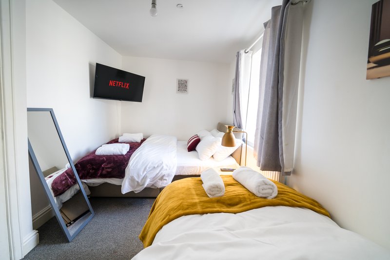 Bright bedroom with two beds, white walls, window with curtains, and wall-mounted TV