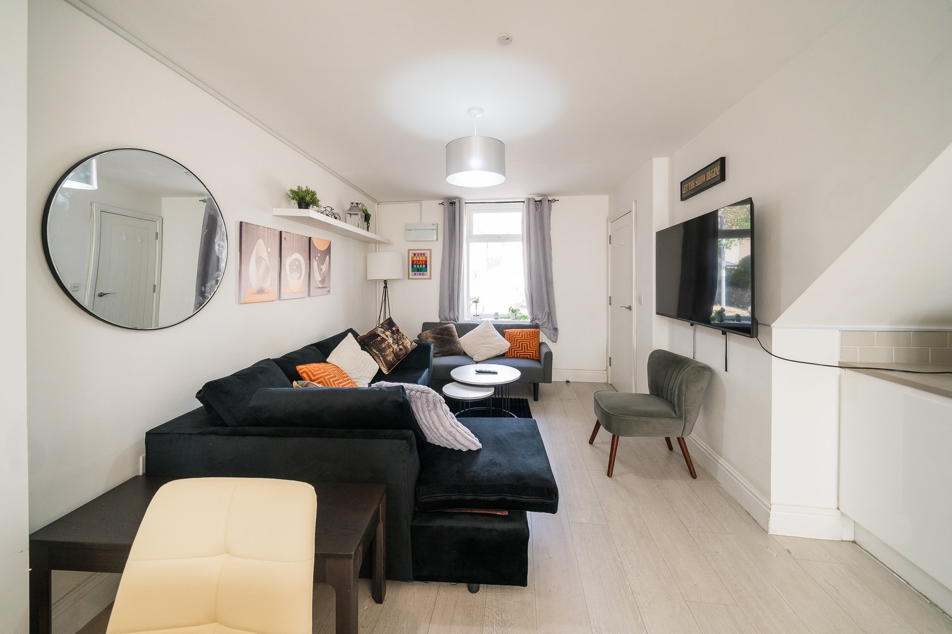 Modern living room with black L-shaped sofa, orange cushions, wall-mounted TV, and round mirror