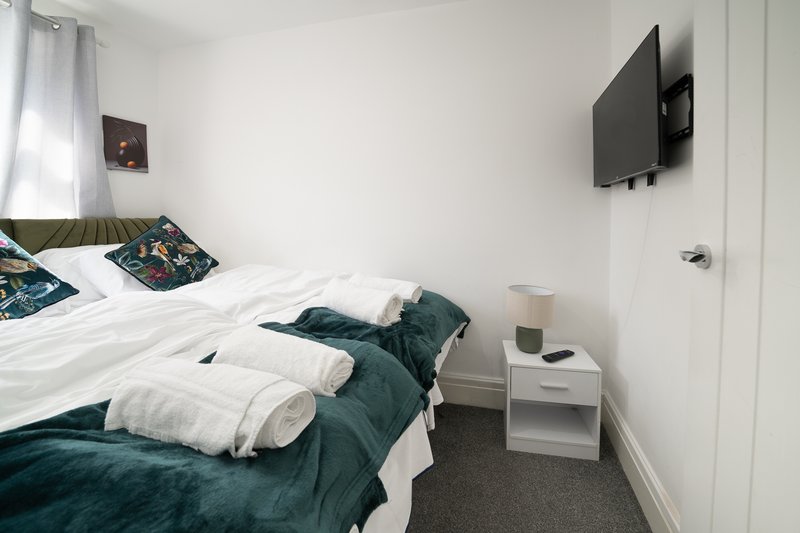 Bedroom with double bed, white bedding, green throw, white nightstand and wall-mounted TV.