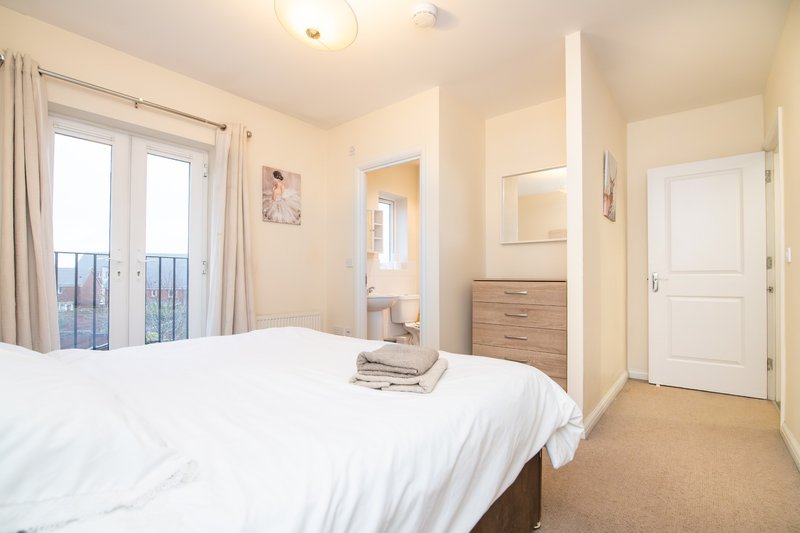 Bright bedroom with white bedding, wooden dresser, ensuite bathroom door, and balcony doors