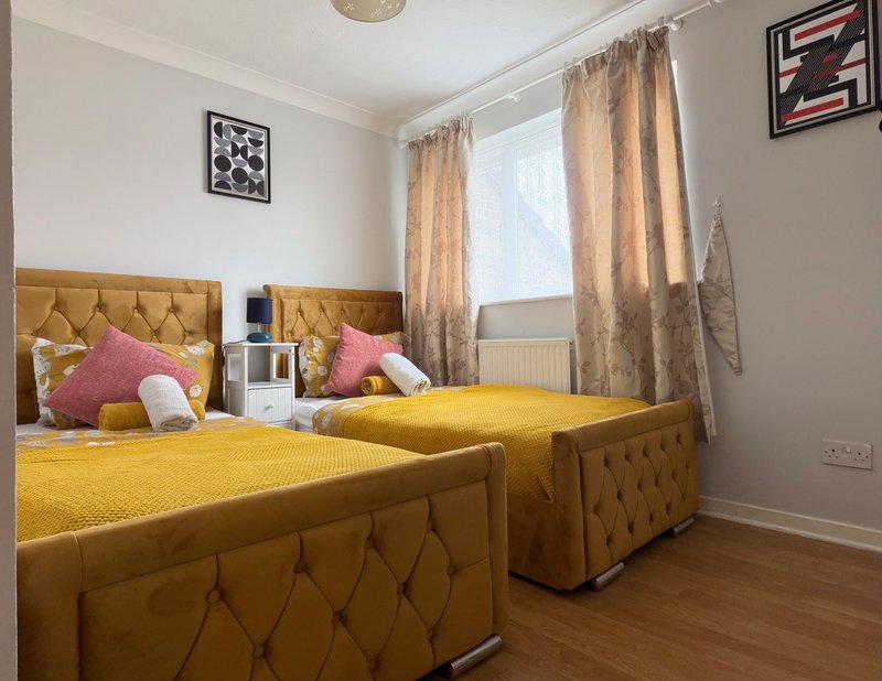 Bright bedroom with two yellow upholstered beds, pink cushions, tan curtains and wooden flooring