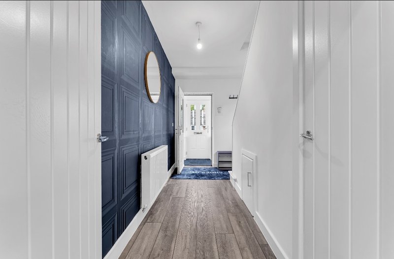 Long hallway with dark blue door, white walls, wooden flooring and circular mirror