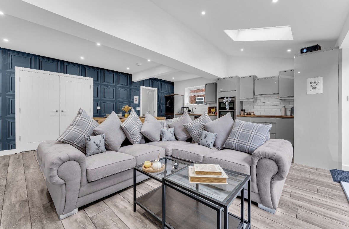 Open-plan living room with grey corner sofa, glass coffee table, and kitchen with dark cabinetry