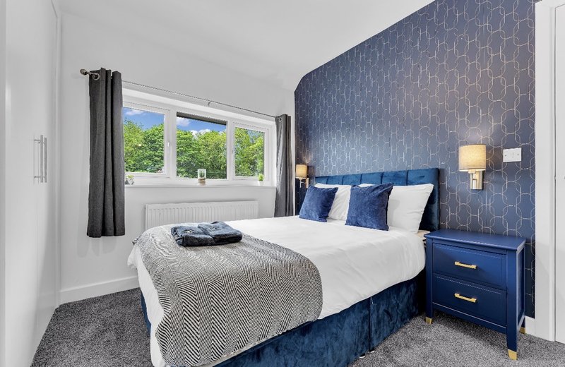 Modern bedroom with double bed, blue headboard, white pillows, and window with garden view