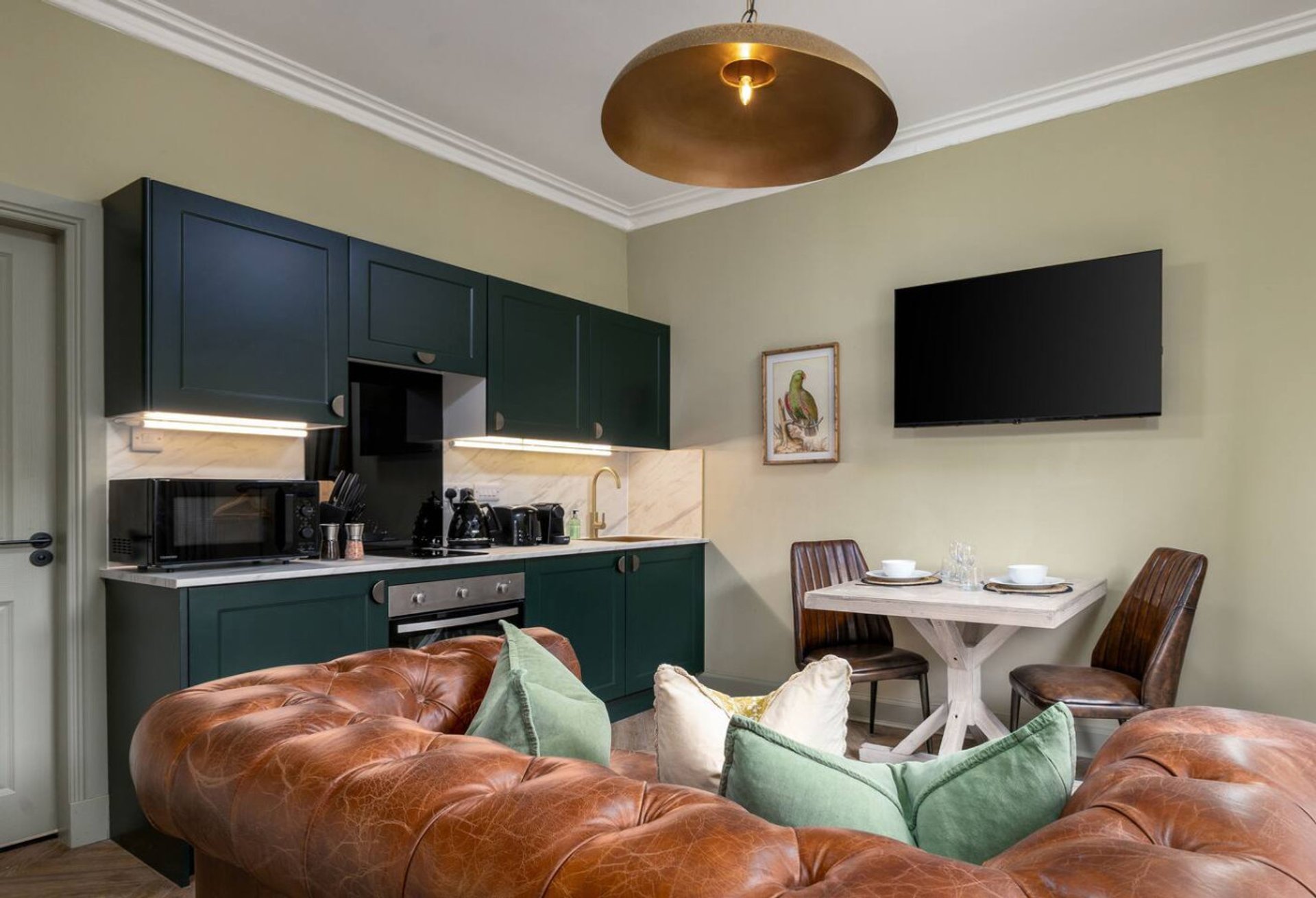 Open-plan kitchen and living area with dark green cabinetry, leather sofa, dining table and pendant lamp