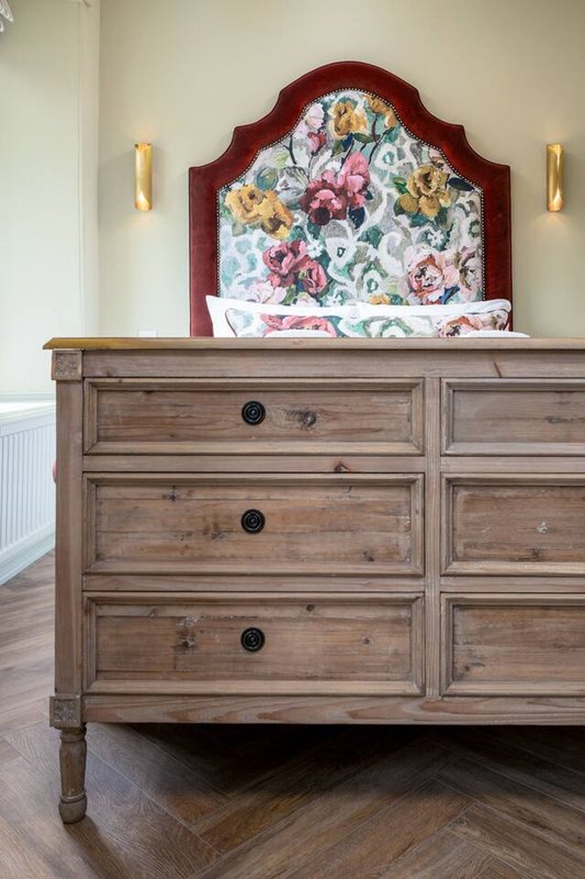 Bedroom with wooden dresser, burgundy upholstered headboard with floral pattern, and gold wall sconces