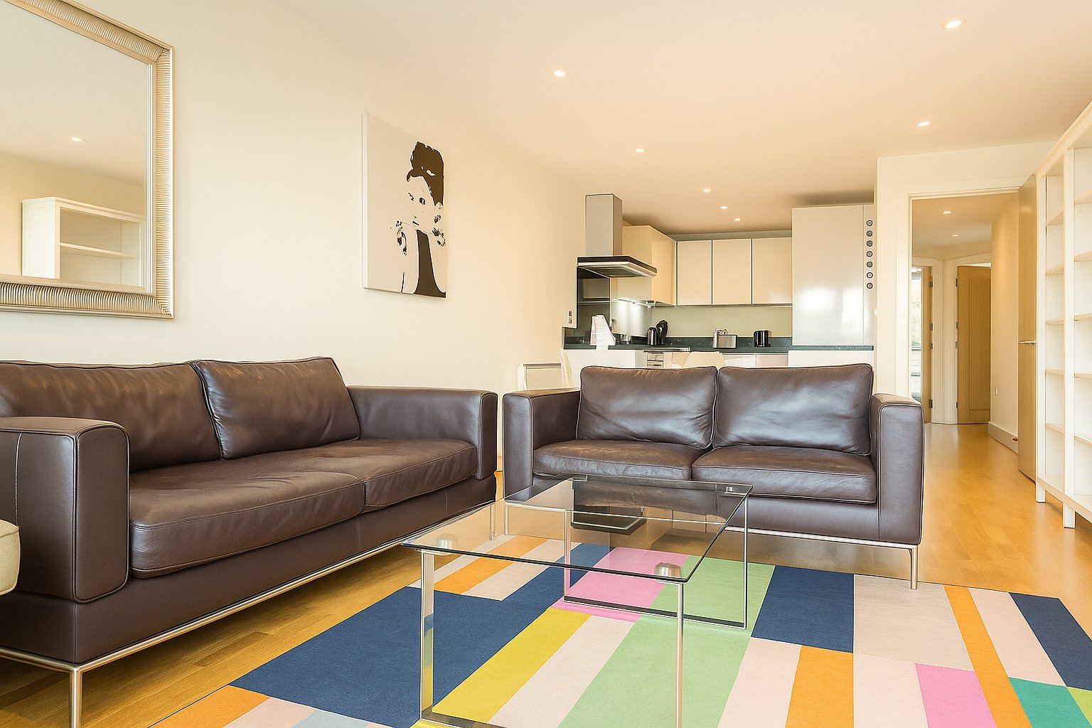 Open-plan living room with two brown leather sofas, colorful geometric rug, glass coffee table and kitchen visible beyond
