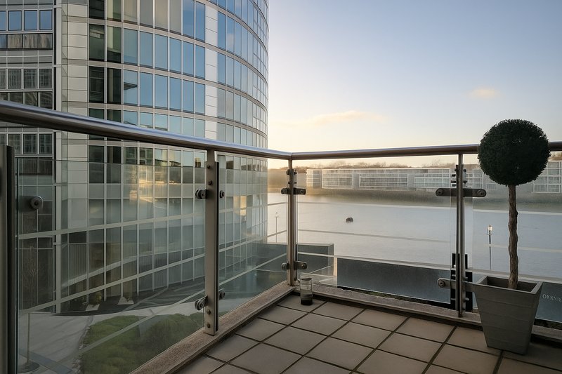 Modern balcony with glass railings overlooking river, contemporary building and potted topiary