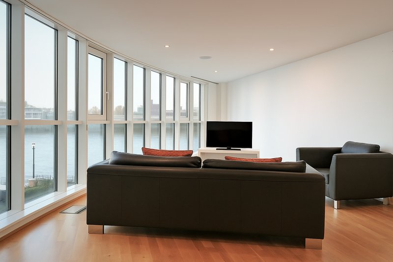 Modern living room with black sofa, TV, large windows overlooking water, wooden floor