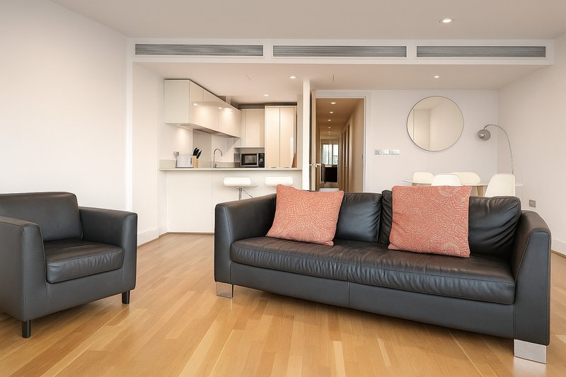 Open-plan living room with black leather sofa, armchair, wooden floor and integrated kitchen area