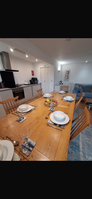 Open-plan kitchen and dining area with wooden table set for four, kitchen appliances, and blue sofa visible in background