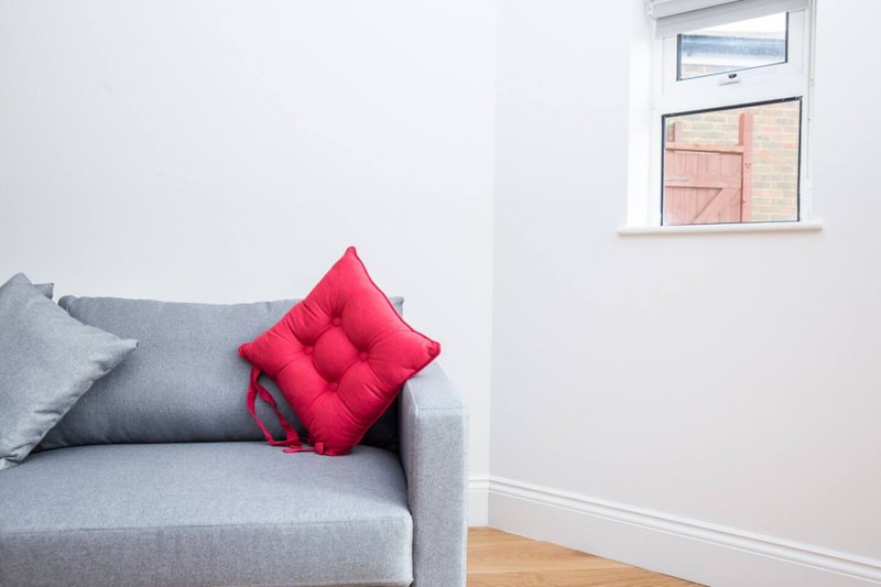 Grey armchair with red and grey cushions, wooden floor, window with brick building view