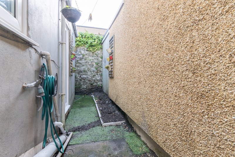 Narrow side passage between two rendered walls with garden beds and coiled hose