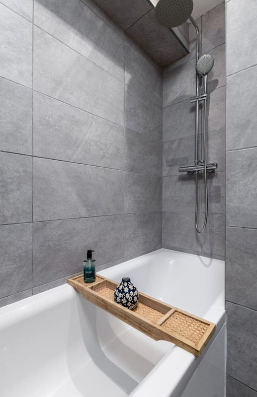 Modern bathroom with white bathtub, grey tile walls, chrome shower head and wooden bath caddy