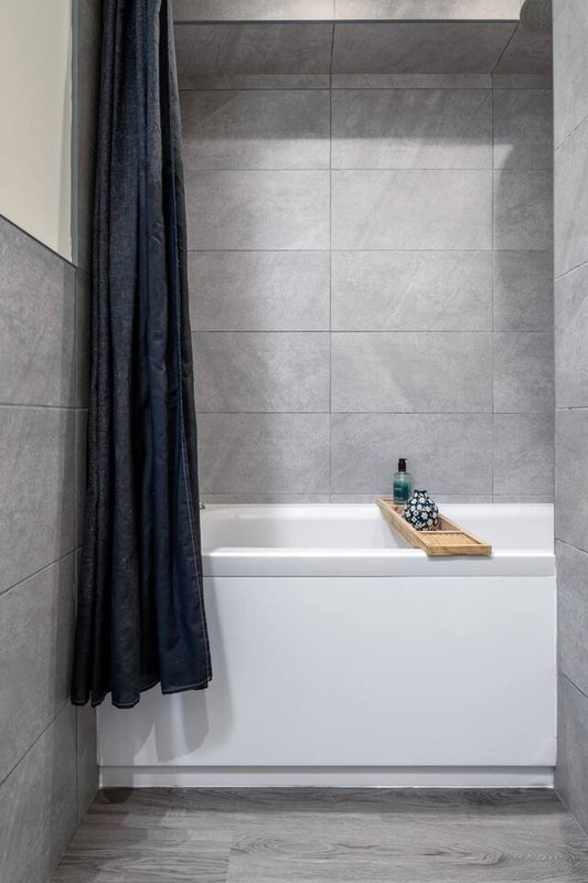 Modern bathroom with gray tile walls, white bathtub, dark shower curtain, and wooden bath caddy
