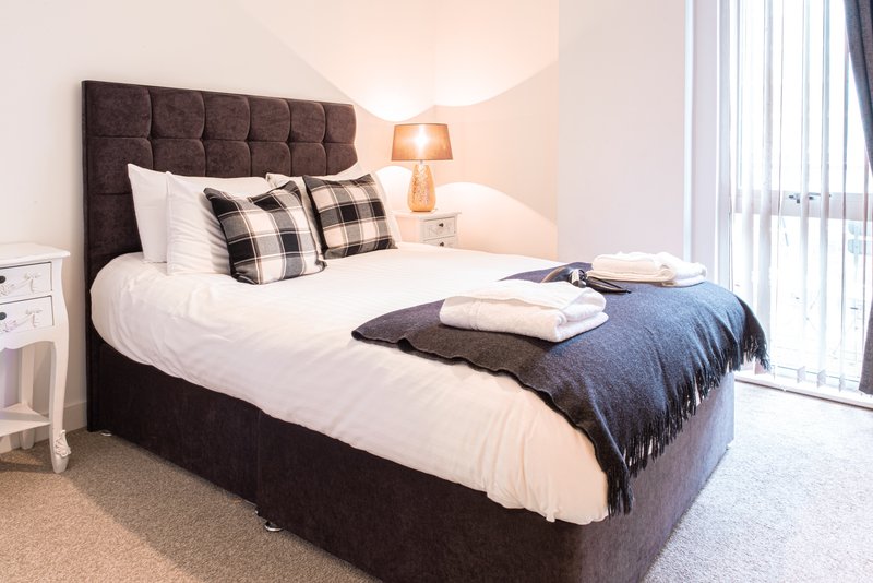 Modern bedroom with dark upholstered bed, white bedding, plaid pillows, and bedside lamp