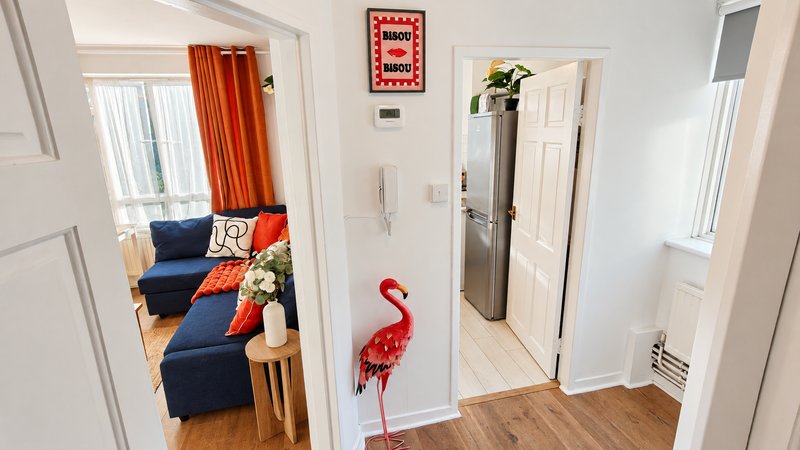 Hallway with white walls, wooden flooring, leading to living room with blue sofa and red curtains